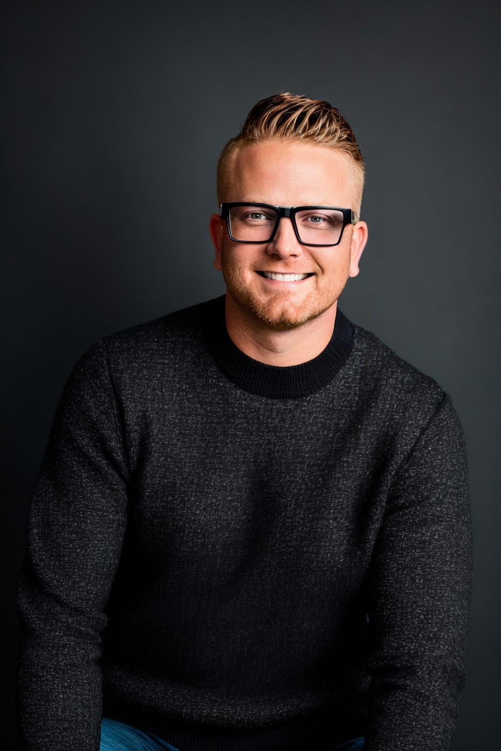 Man in glasses smiling, gray sweater, professional portrait.